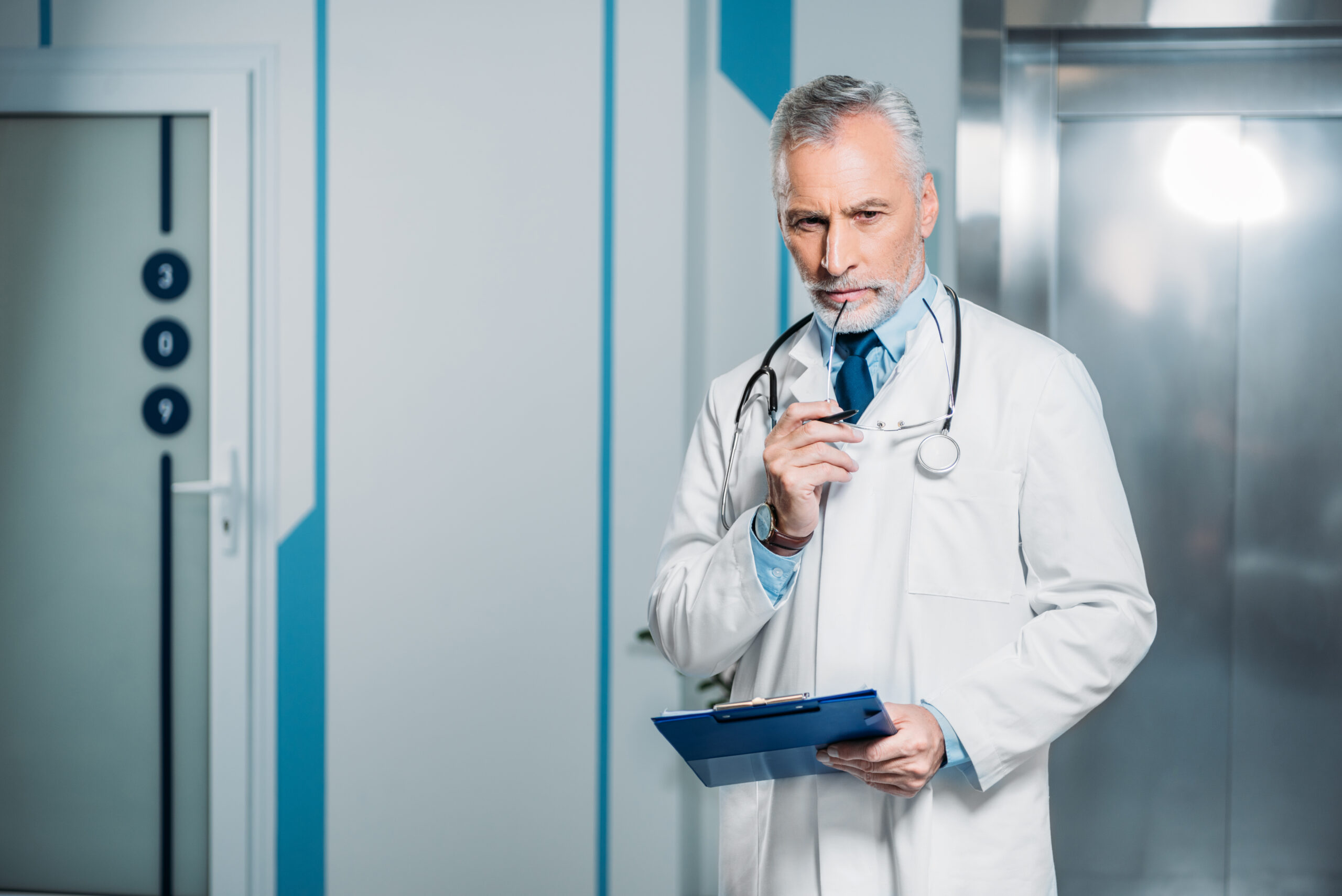 Pensive Mature Male Doctor Holding Eyeglasses And Clipboard In Hospital - ARBOS CONTABILIDADE